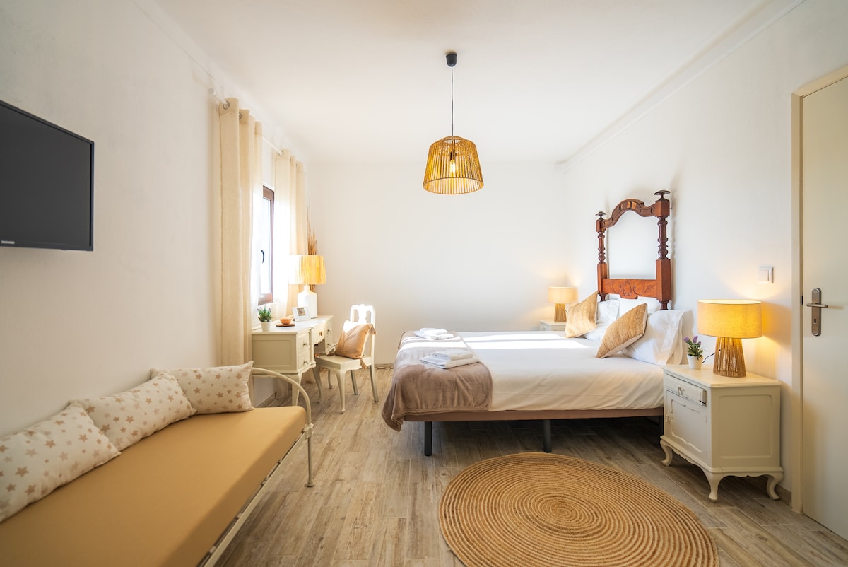 The suite features two individual beds and a small bed suitable for a child. A seating area is presented with a sofa and a side table. Natural light fills the room, highlighted by soft curtains and a woven rug underfoot. Decorative lamps add a warm glow.