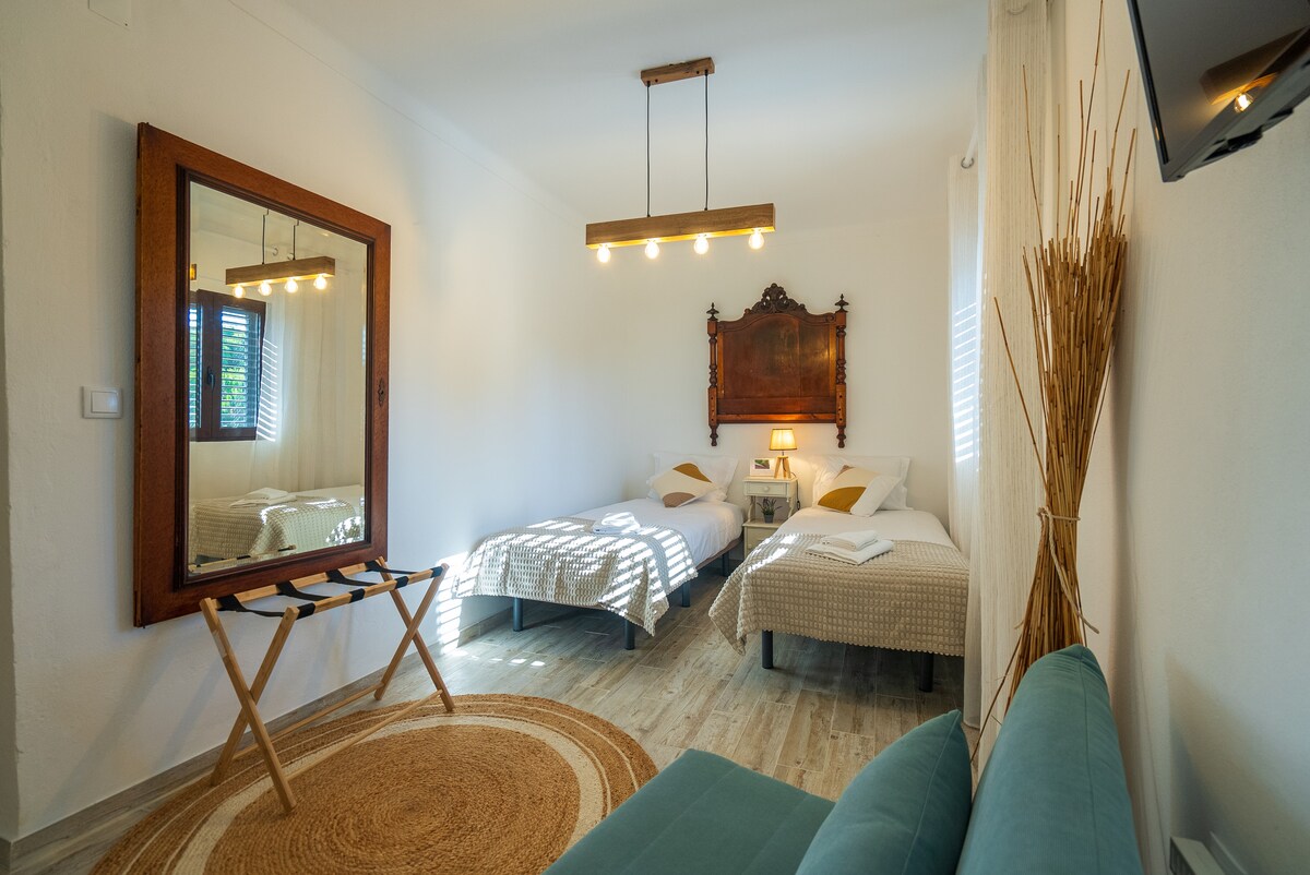 A bright sleeping area features two individual beds with neutral bedding and decorative pillows. A large mirror reflects natural light, while a cozy rug is positioned on the wooden flooring. A stylish seating area includes a small sofa, complementing the overall inviting ambiance.