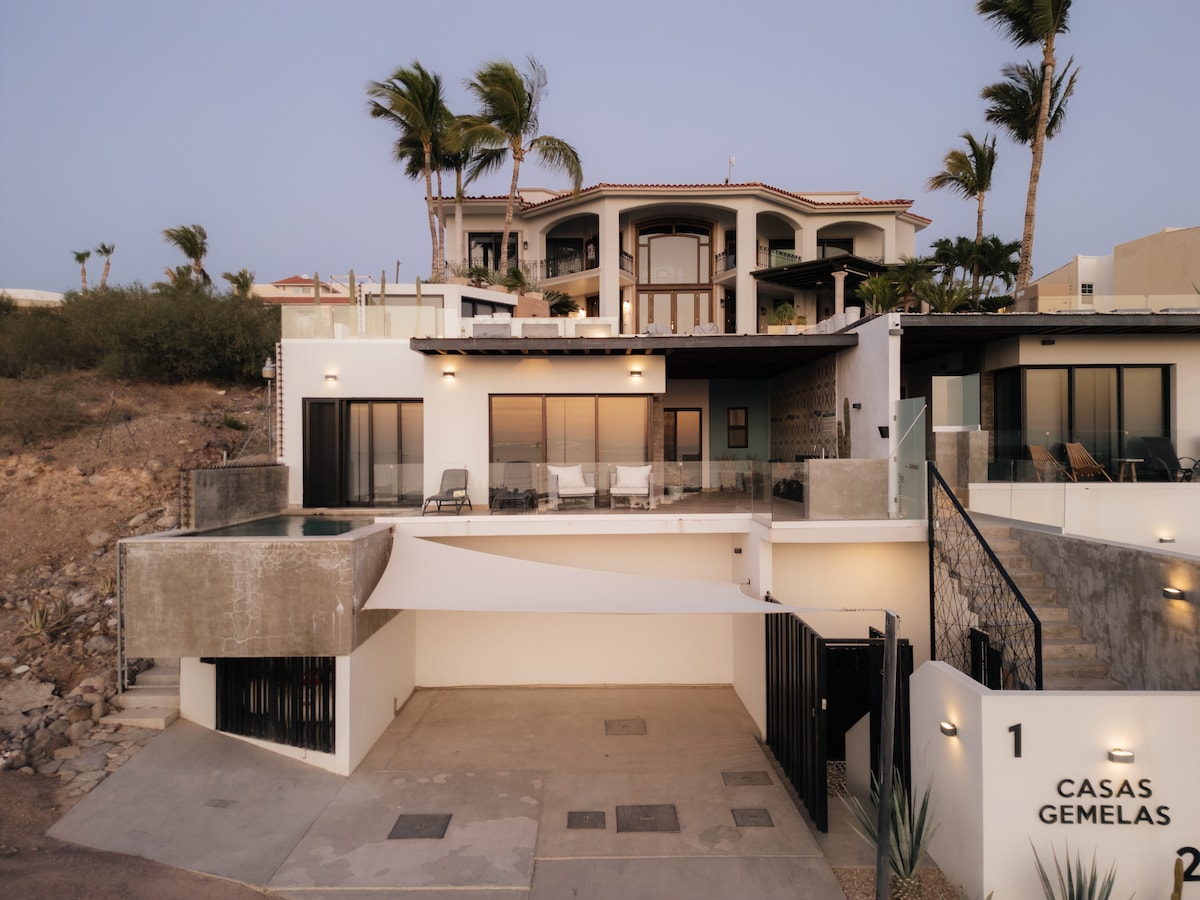 A modern multi-story home with a sleek exterior is shown. The private pool and spacious terraces are visible, surrounded by lush landscaping. The entrance is framed by a minimalist design, featuring stylish gates and clear views of the surrounding area, with the sunset casting a soft glow.