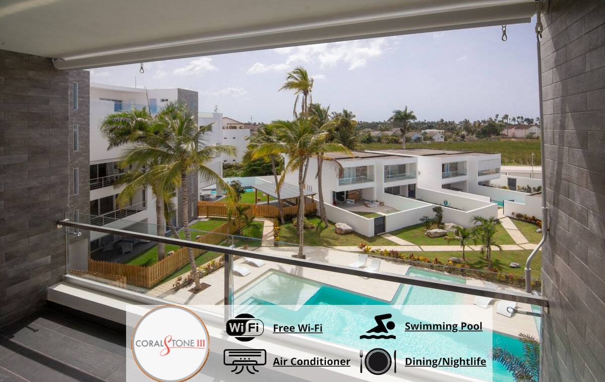 A view from the terrace showcases the swimming pool below, surrounded by palm trees and landscaped gardens. Modern buildings are visible in the background, contributing to the ambiance of the outdoor space. The terrace features glass railings, allowing for unobstructed views.