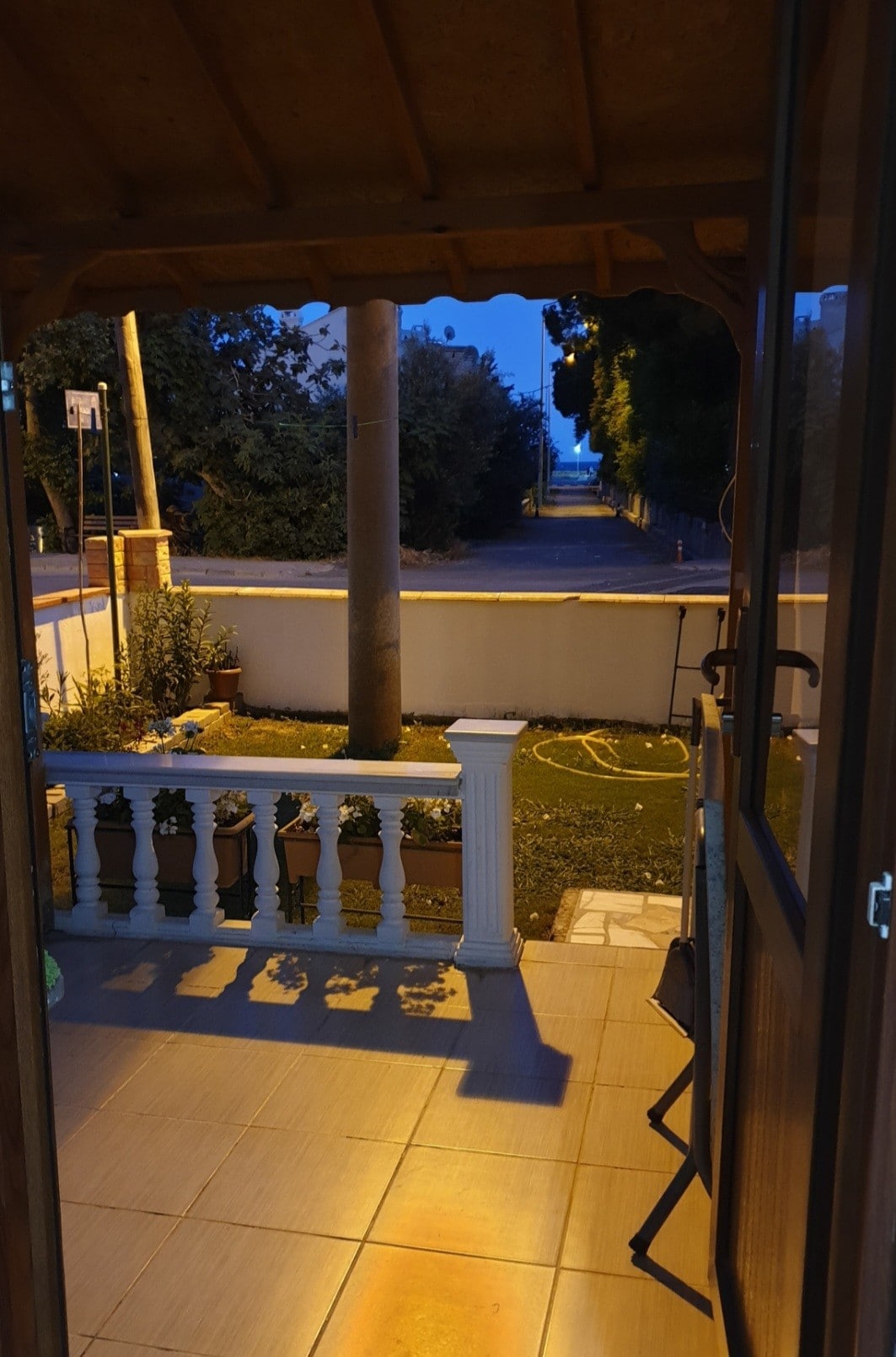 A porch is visible in the forefront, leading to a pathway flanked by trees and distant buildings. The ground is tiled and includes a small garden area, with planter boxes placed along the railing. Soft evening light frames the view, creating a serene ambiance.