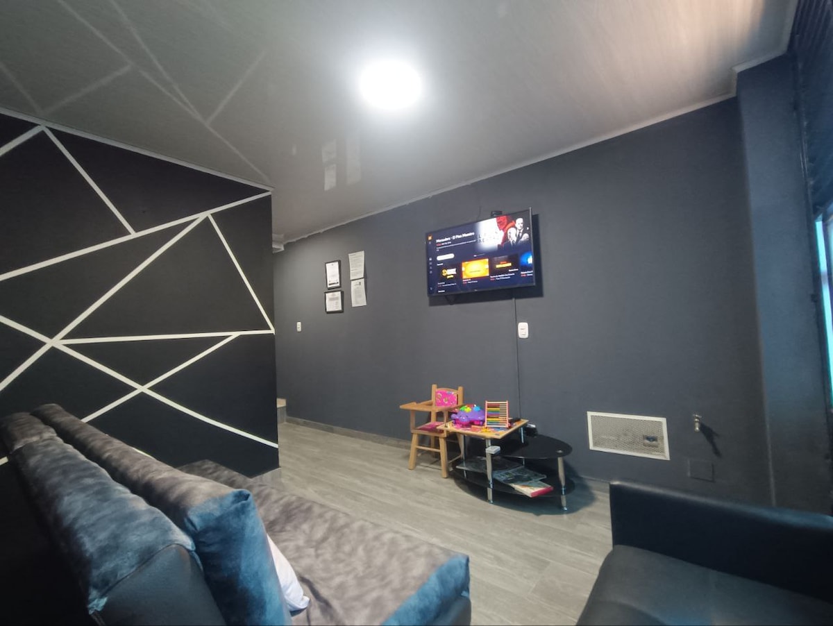 A modern living room features a dark accent wall with geometric designs. A flat-screen television is mounted on the wall, displaying content. A comfortable sofa is arranged in a casual setting, accompanied by a small table with toys, enhancing a welcoming space.