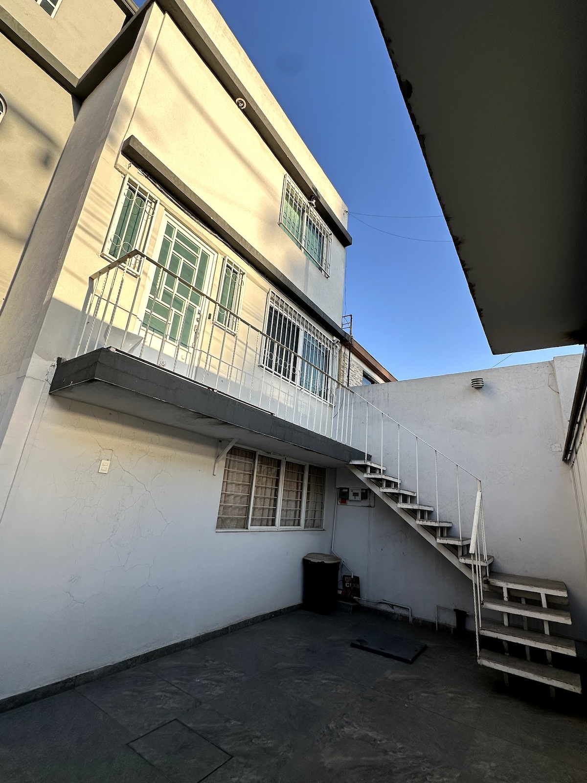 The exterior features a multi-story building with a staircase leading to the upper level. Metal railings surround the stairs, and large windows allow natural light into the structure. The courtyard area is paved with stone, providing a clear and open space.