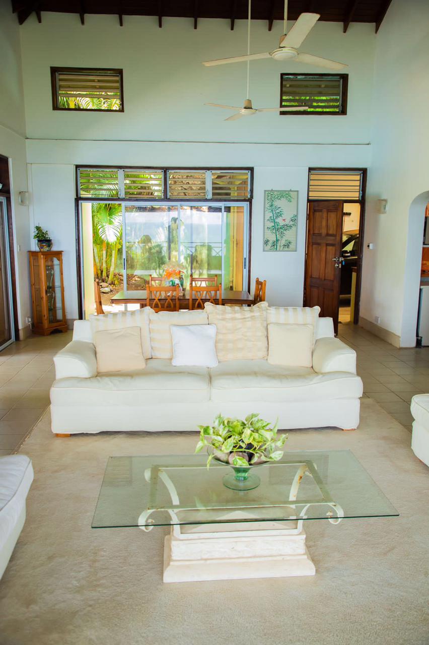A spacious living area features light-colored sofas and a glass coffee table. Natural light floods the room through large windows, showcasing views of the outdoor dining space. Tropical plants are visible, adding a touch of greenery to the inviting environment.