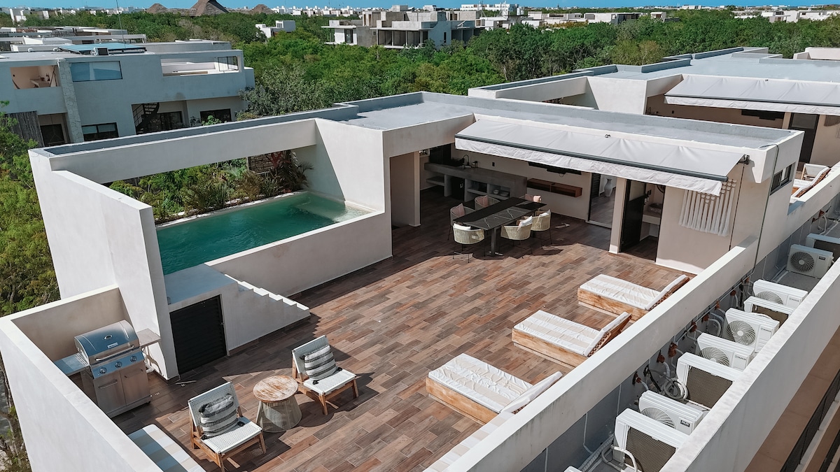 The rooftop terrace features a well-equipped outdoor kitchen and a spacious dining area. Two heated pools are visible, surrounded by comfortable lounge chairs. The area is designed for relaxation, with a blend of greenery in the background creating a serene environment.