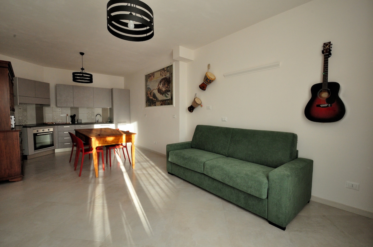 A modern living space features a green sofa facing a wooden dining table surrounded by red chairs. A kitchen area is visible with gray cabinets and stainless steel appliances. Decorative items, including a guitar and artworks, add character to the light-filled room.