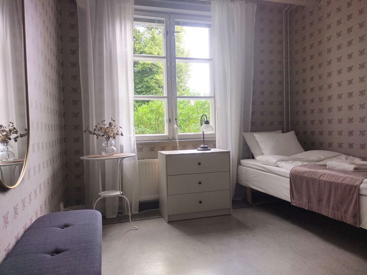 A simple and serene guest room features a spacious bed, soft linens, and a bedside table with a lamp. Natural light enters through a large window, which is framed by light curtains. A decorative mirror hangs on the wall, and a small table holds dried flowers.