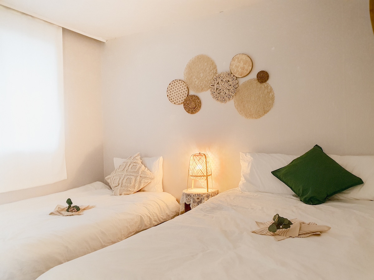A serene sleeping area is featured, showcasing two spacious beds adorned with neutral bedding. A decorative lamp emits a gentle glow beside one bed, complemented by a collection of woven wall hangings. Natural light filters through a window, enhancing the calming atmosphere.