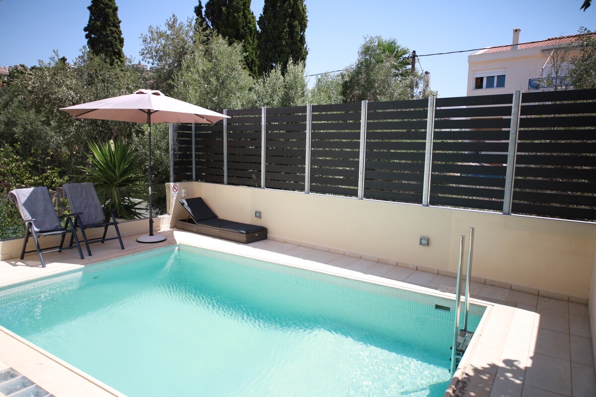 A private pool area is visible, complete with a light blue pool and surrounding greenery. Two lounge chairs and a sunbed are positioned beside the pool, while an umbrella provides shade. The modern fencing offers privacy and enhances the serene atmosphere.