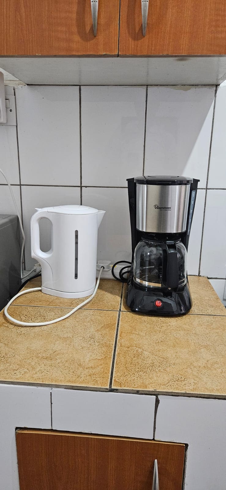 A kitchen counter is shown with a white electric kettle and a black coffee maker. Both appliances are positioned next to each other against a tiled backsplash, with the counter surface featuring a beige and brown design. The setting offers a functional space for meal preparation.