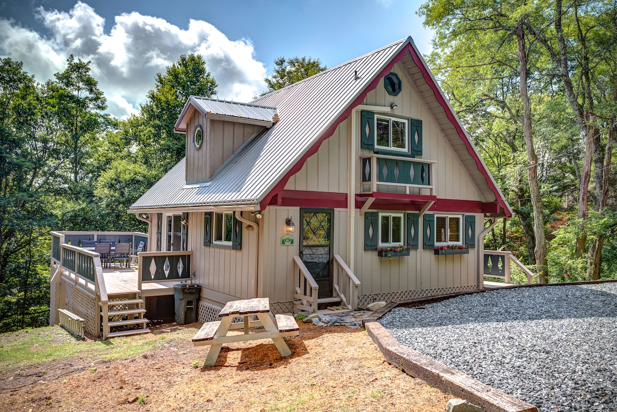 The exterior of the chalet features a unique architectural design with colorful accents. A spacious wrap-around porch is visible, accompanied by outdoor seating options. Surrounded by trees, the setting offers a serene atmosphere, while a gravel area provides convenient access to the entrance.