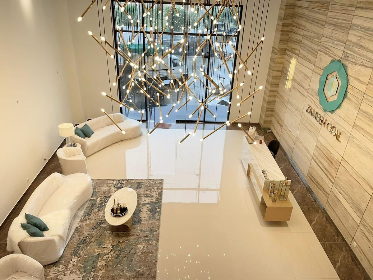 A spacious lobby area features modern seating arrangements, with a large light fixture creating a focal point above. The polished floor reflects natural light coming through expansive windows, and a reception desk is positioned beside a decorative wall with the property logo.
