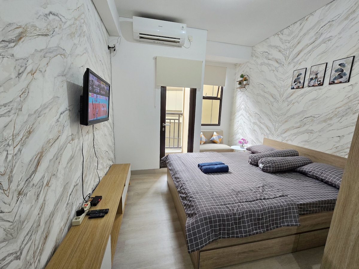 The studio bedroom features a spacious bed adorned with neutral-colored bedding. A television is mounted on the wall across from the bed. Natural light enters through a window, complemented by decorative wall art and a small potted plant on a shelf.