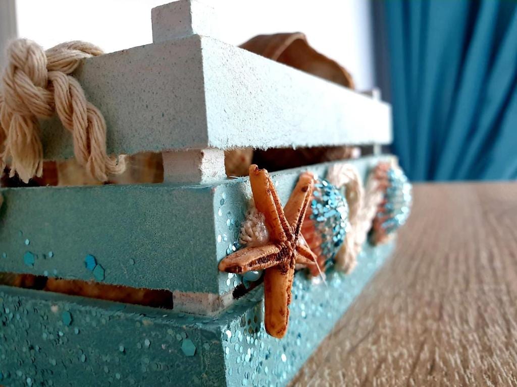 A decorative wooden box in ocean-inspired colors features a textured surface adorned with starfish and shells. The box is positioned on a textured wooden table, with light reflecting off its turquoise finish, complemented by a soft background in varying shades of blue.