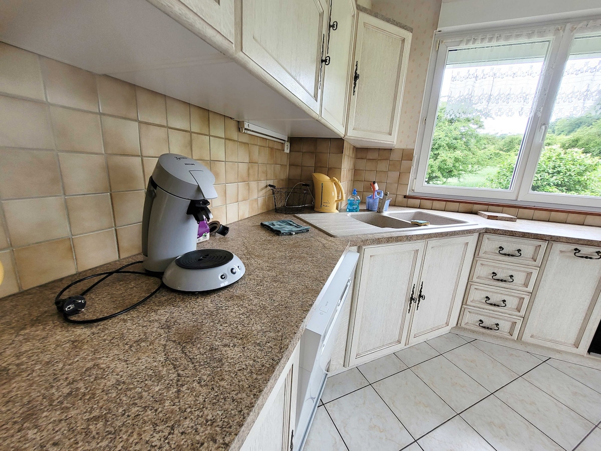 The kitchen features light-colored cabinetry with intricate hardware, complemented by a spacious countertop. A coffee maker and kettle are positioned on the counter, while a window allows natural light to illuminate the area, revealing a view of the greenery outside.