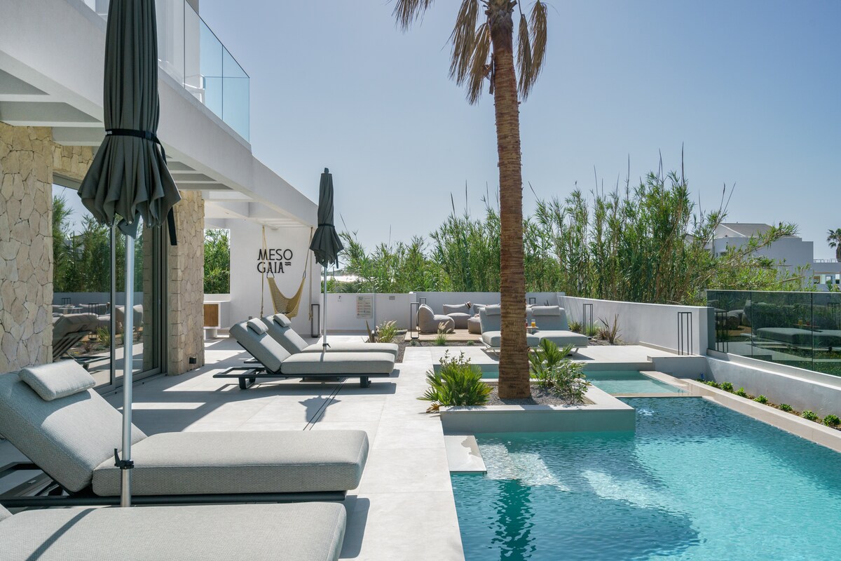 A spacious outdoor area features a heated swimming pool surrounded by lounge chairs under shaded umbrellas. Lush greenery adds a natural touch, while a stone wall displays the Mesogaia Villa sign. Sun loungers invite relaxation beside the calm water.