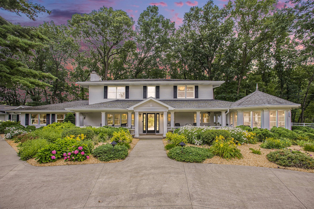 The welcoming exterior of the retreat is framed by lush greenery and vibrant flower beds. A spacious driveway leads to the main entrance, which features large windows and a prominent porch. The serene surroundings and evening sky enhance the inviting appearance of the home.