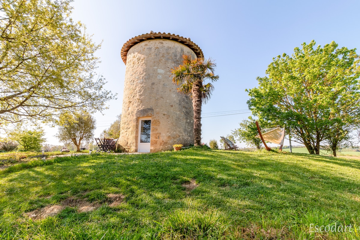 The stone structure, resembling a mill, is set amidst a lush green landscape. Surrounding trees provide shade, while a hammock and outdoor seating area are accessible in front. A clear blue sky creates a serene atmosphere for relaxation in nature.