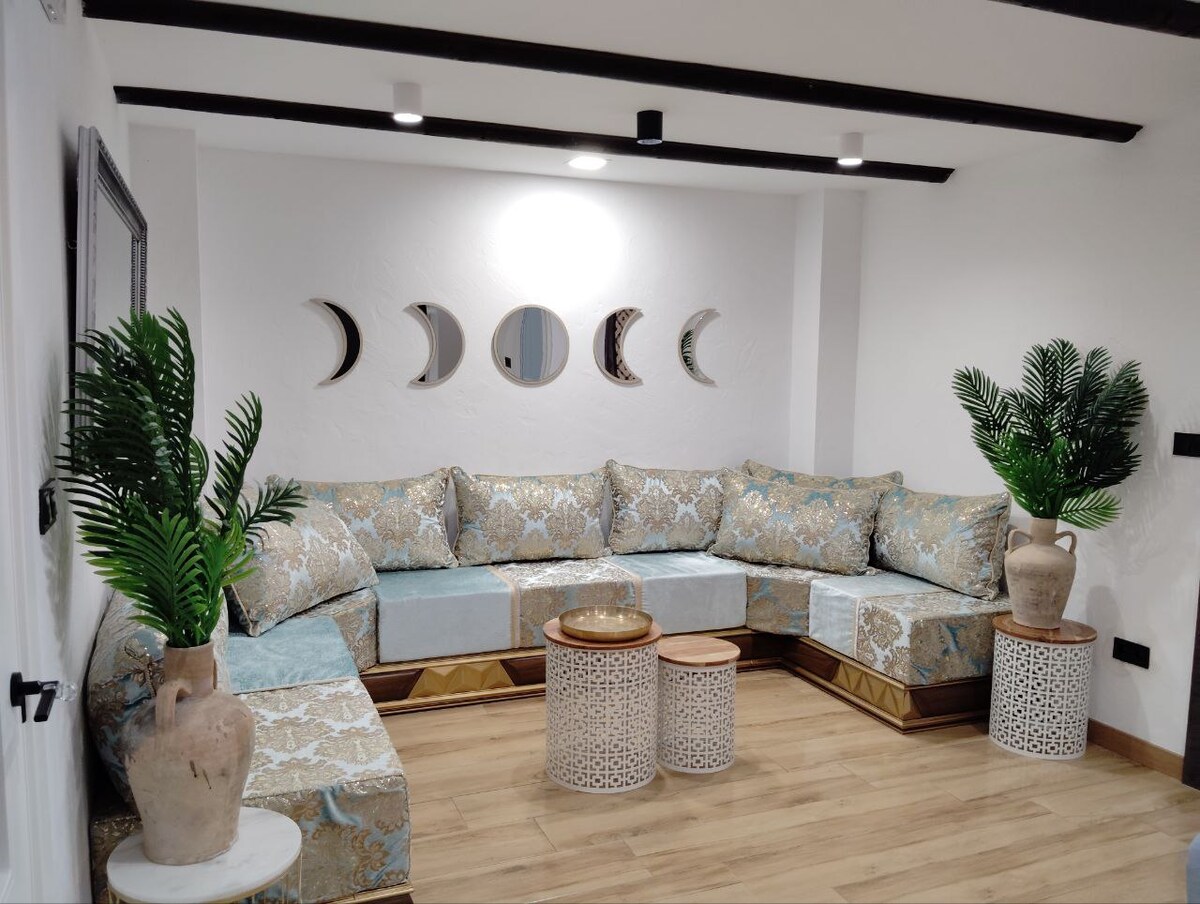 A spacious living area features a U-shaped seating arrangement with patterned cushions in soft tones. Decorative tables made of textured materials are positioned in front of the seating. Greenery is provided by two potted plants, while decorative mirrors are mounted on the wall.