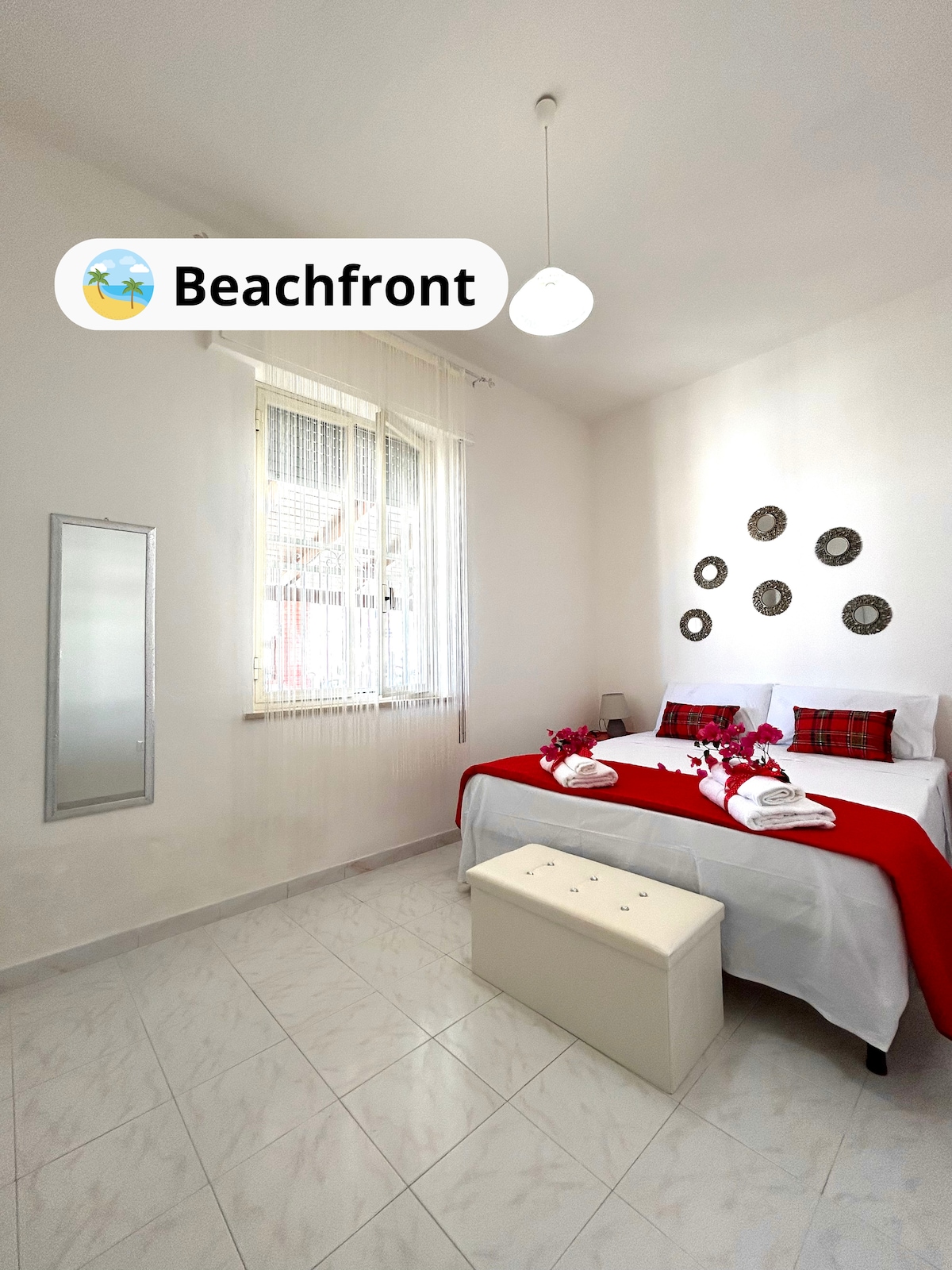 A well-lit bedroom features a spacious bed with red and white bedding. Decorative mirrors hang on the wall, complementing the light and airy atmosphere. A small bench sits at the foot of the bed, and natural light filters in through the window adorned with curtains.