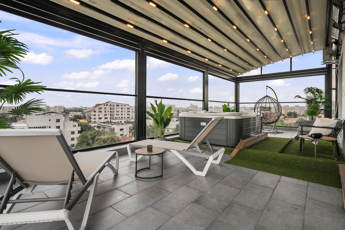 An expansive terrace features a relaxing spa tub and comfortable seating areas. Abundant natural light floods the space through large windows, showcasing panoramic city views. The area is adorned with greenery and ambient lighting, creating a serene environment ideal for relaxation or gatherings.