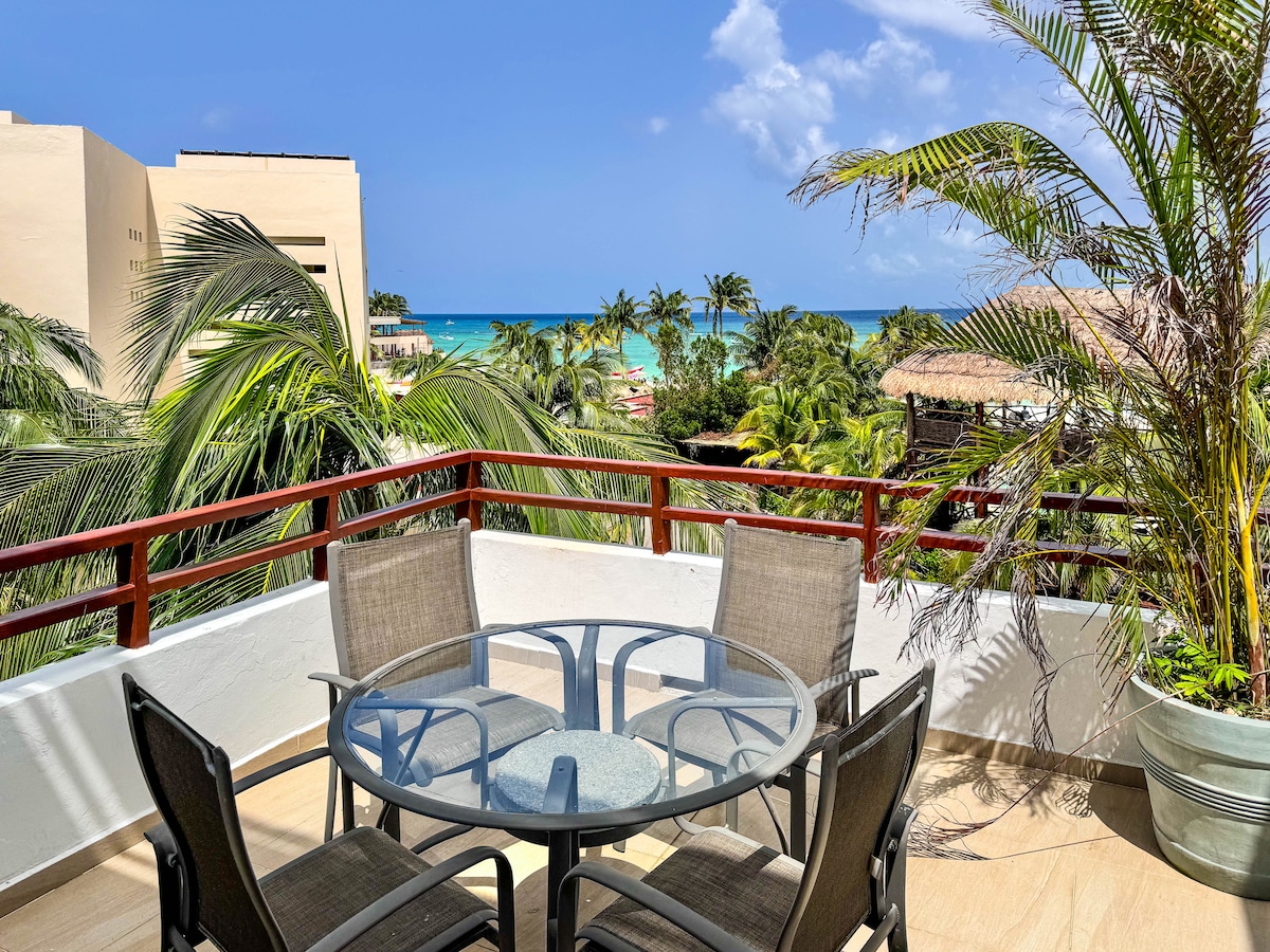 A spacious ocean-view patio is featured, furnished with a round glass table surrounded by four comfortable chairs. Lush palm trees frame the scene, while the tranquil ocean can be seen in the background, enhancing the inviting outdoor space.