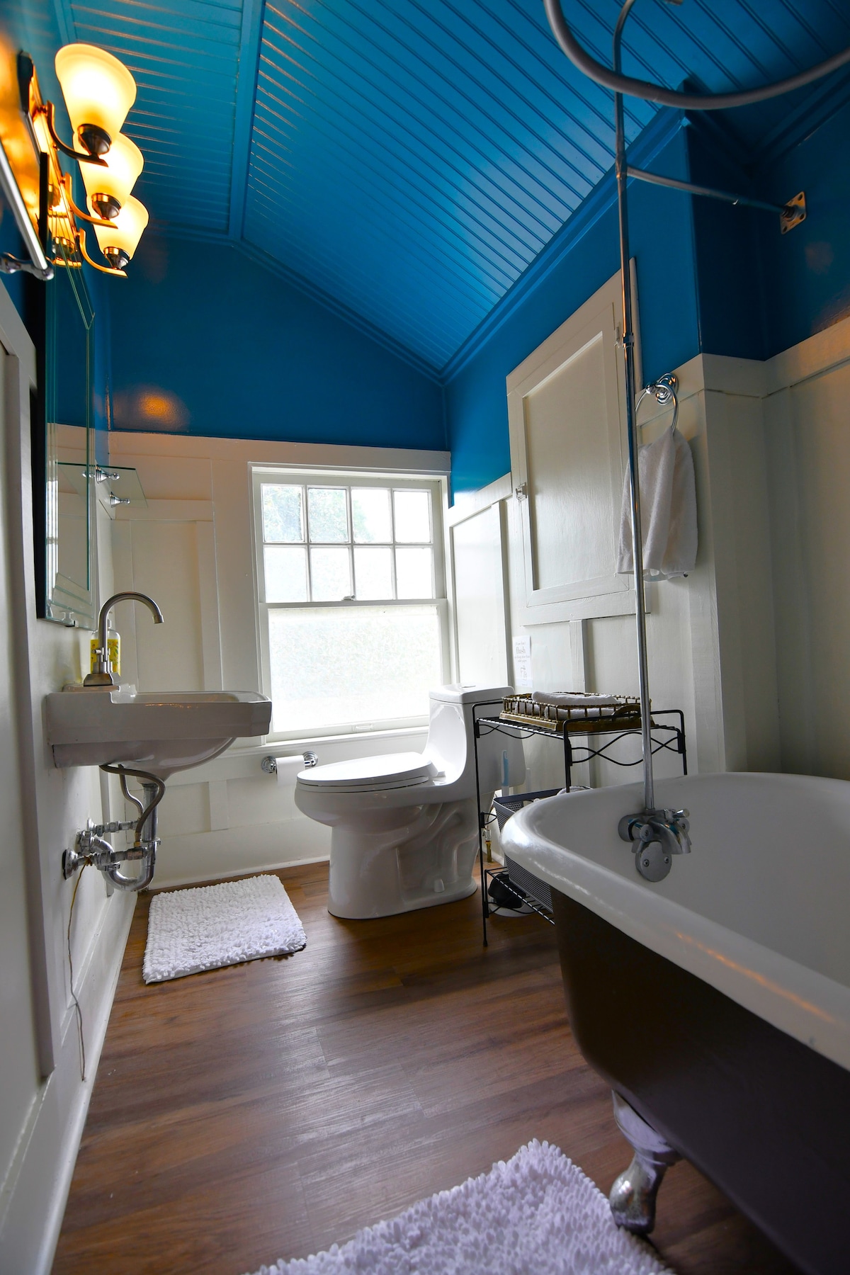 The bathroom features an antique clawfoot tub and a modern toilet. A wall-mounted sink is situated near a well-lit mirror. Bright blue walls are complemented by light wood flooring and white accents, creating a refreshing atmosphere. Natural light enters from a window with frosted glass.