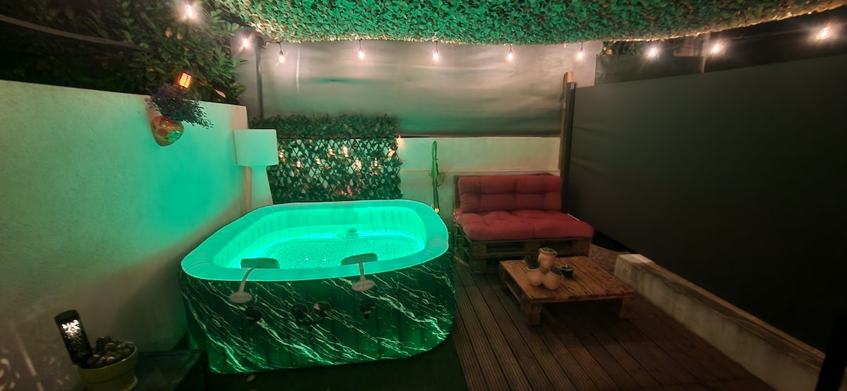 A private outdoor spa is illuminated with soft green lighting, inviting relaxation. A cozy seating area with a plush couch is present nearby, complemented by a wooden pallet table. Soft overhead lights enhance the tranquil ambiance, surrounded by natural foliage.