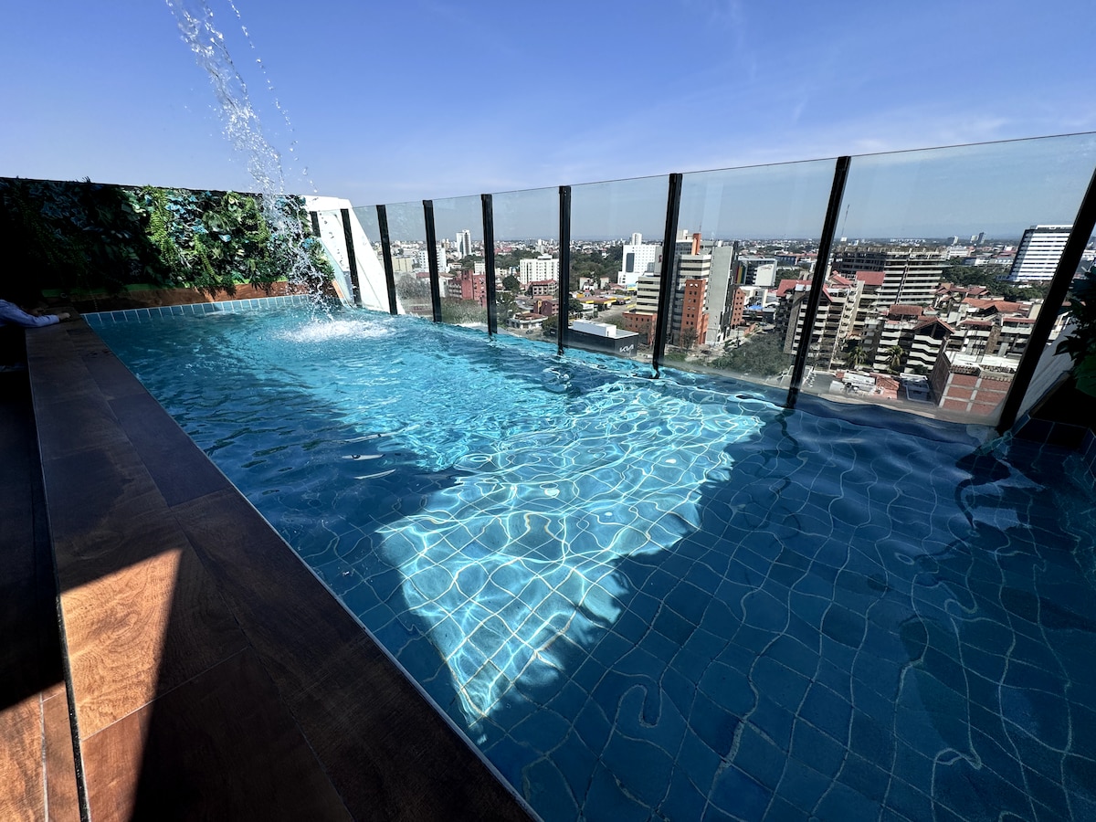 A rooftop pool is showcased with clear blue water, offering views of a vibrant city skyline. The pool's edge features glass panels, enhancing the sense of openness. Sunlight creates reflections on the water's surface, while green foliage lines one side, providing a natural touch.