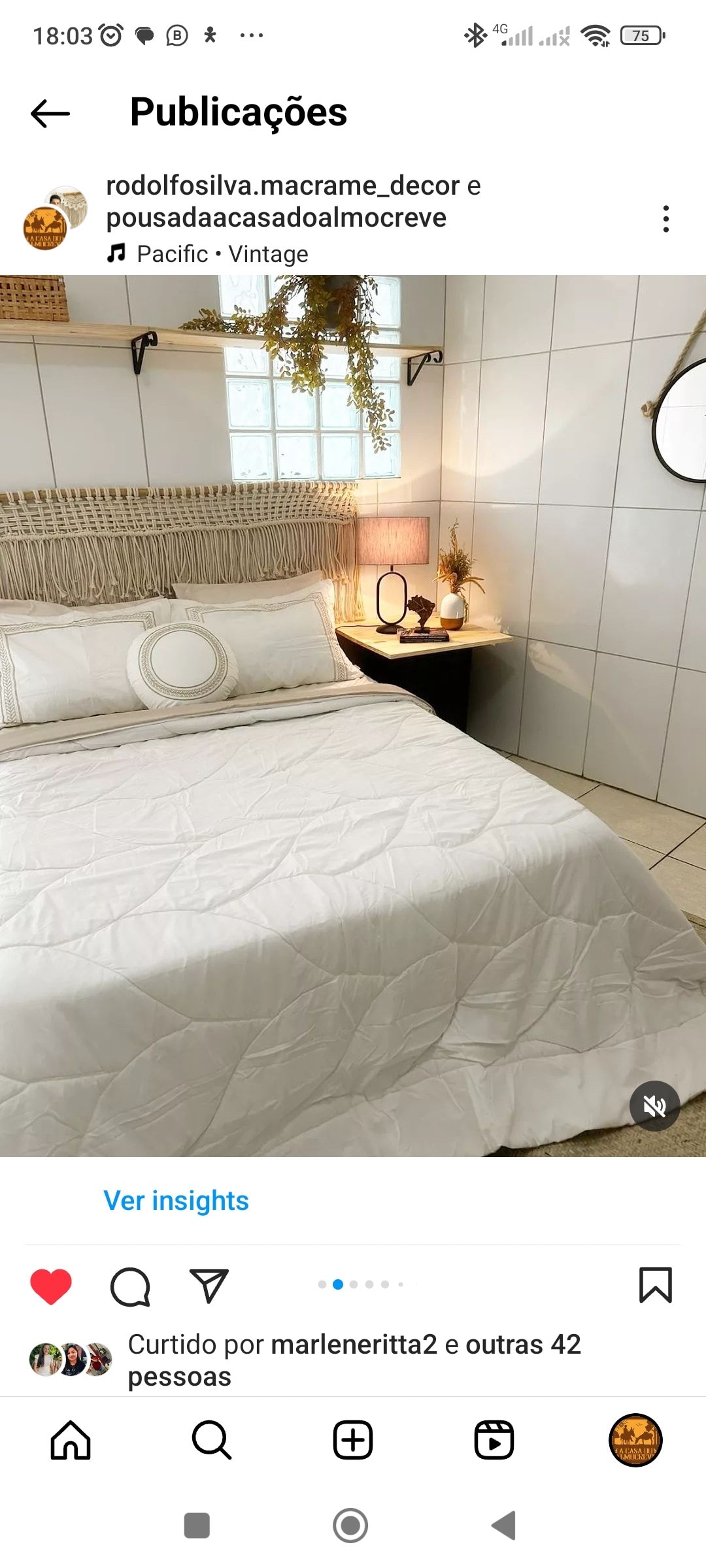 A cozy sleeping area features a light-colored quilted bedspread draped over a spacious bed. A round decorative pillow is placed against the headboard. A bedside table hosts a lamp and a small plant, while a window allows natural light to brighten the room.