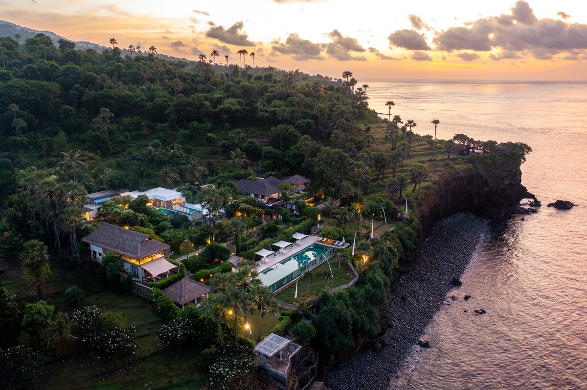 An aerial view captures the resort's lush landscape, with various low-rise buildings nestled among greenery. The coastline is visible, along with an infinity pool that overlooks the ocean, creating a serene atmosphere as the sun sets in the background.