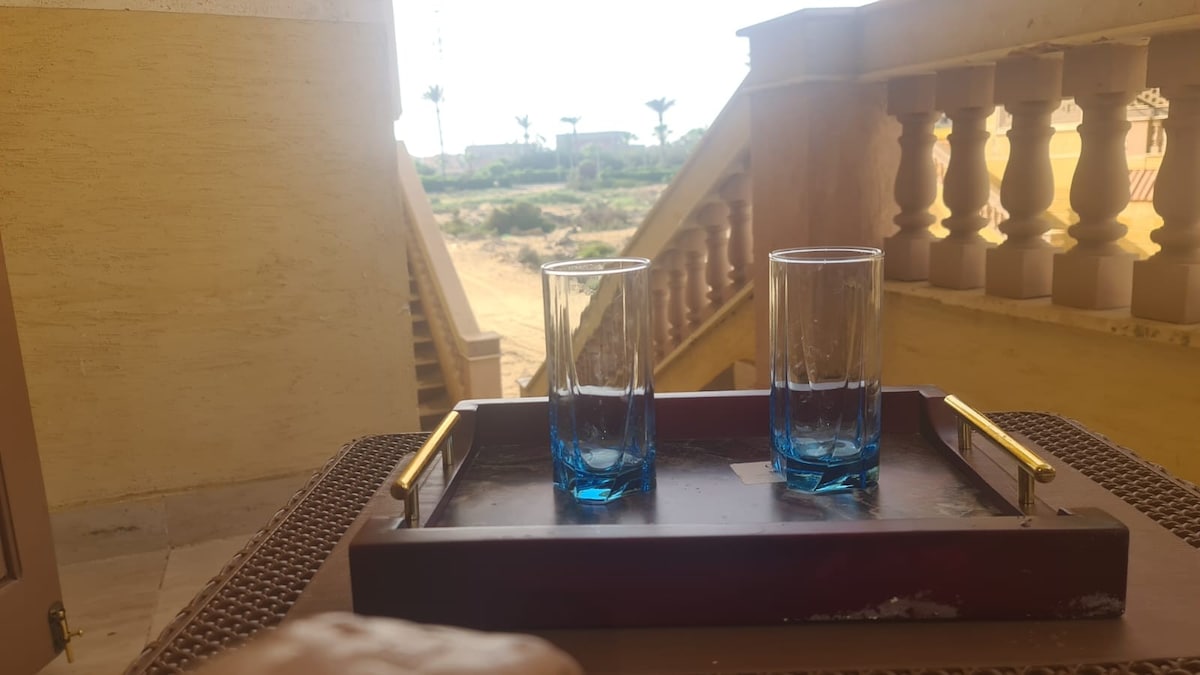 A wooden tray is positioned on a surface, featuring two clear glasses with blue bases. In the background, a staircase leads to an outdoor area with palm trees and a view of open land under a bright sky.