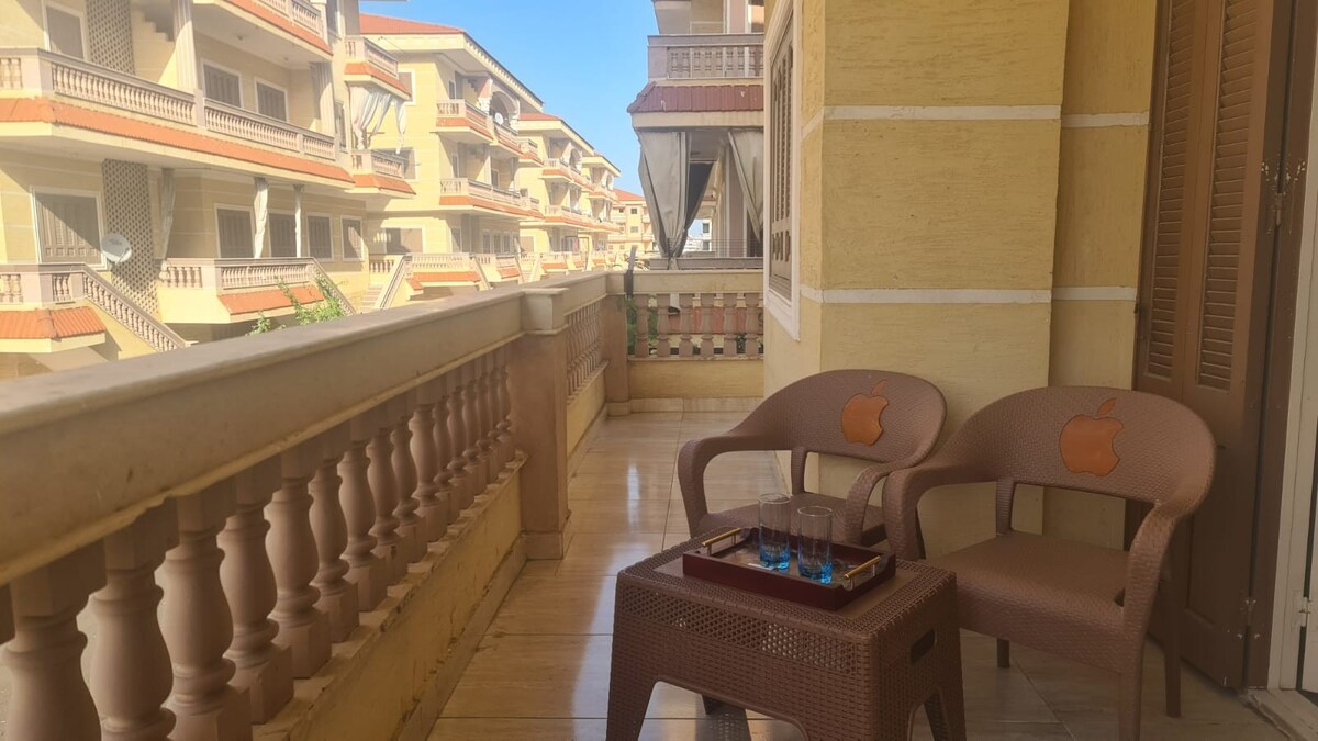 A sunlit balcony features two wicker chairs with armrests and a small table, ideal for relaxed seating. The surrounding architecture includes multiple buildings with balconies, creating a sense of openness and connection to the outdoors.