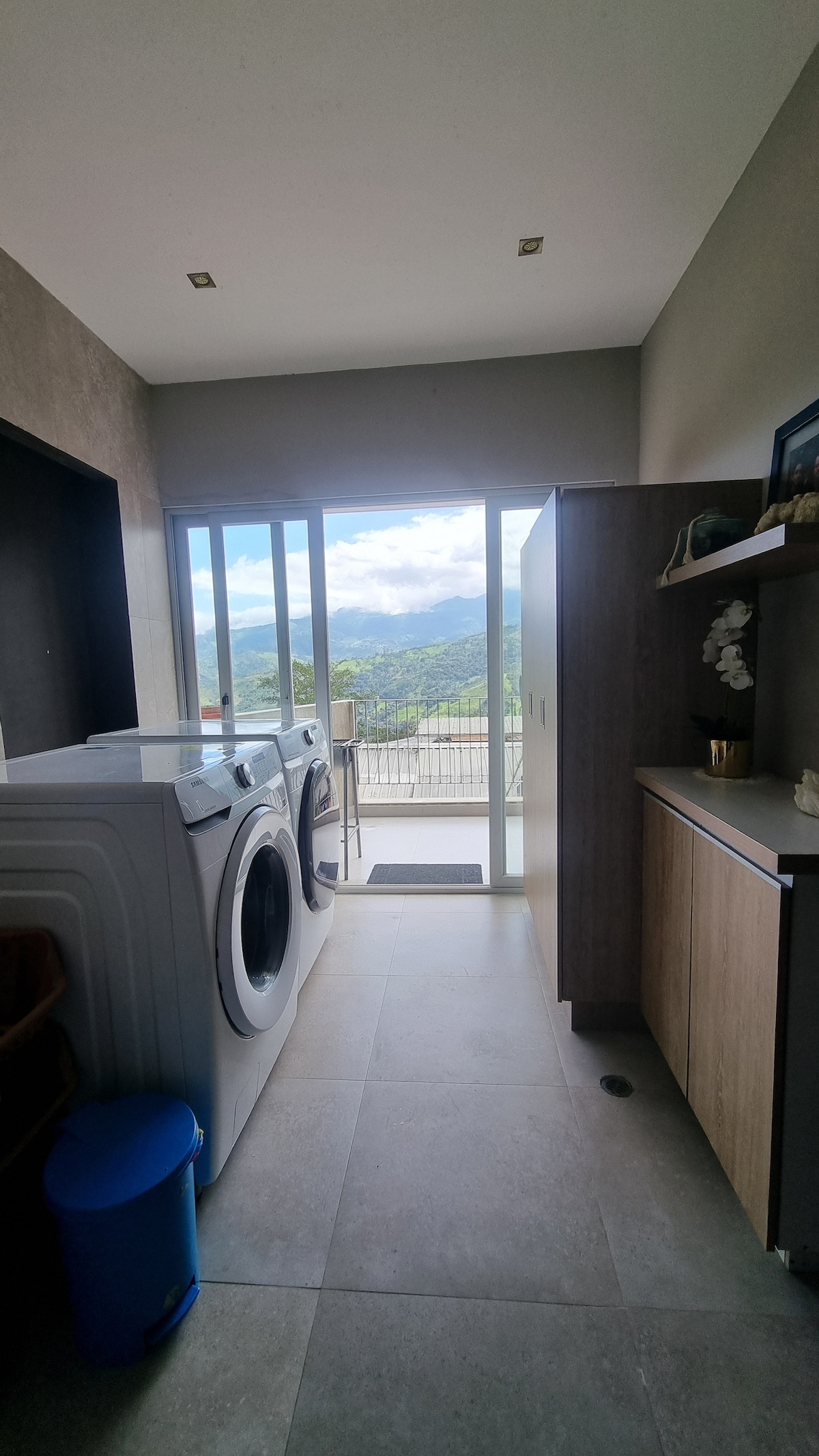 A well-lit laundry area is equipped with a washing machine and dryer, positioned beside a sleek storage cabinet. Large glass doors provide a view of the surrounding mountains, allowing natural light to fill the space.