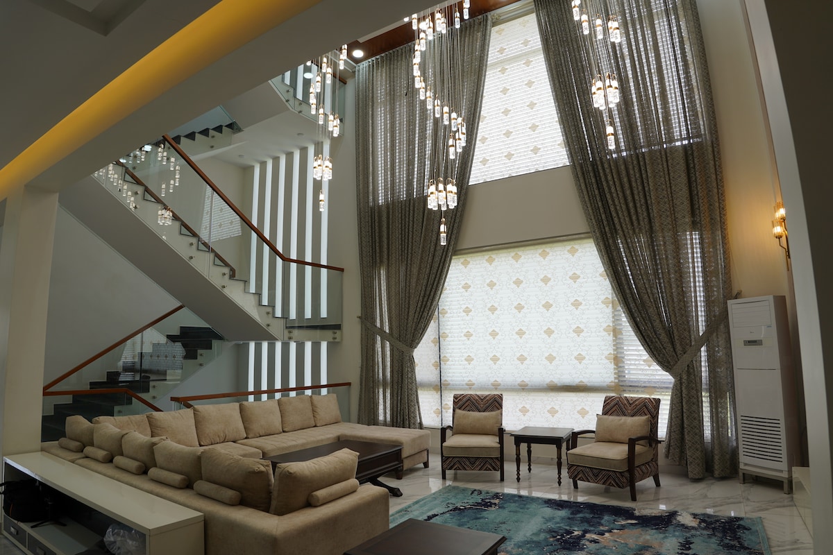 A spacious living area is highlighted by tall windows adorned with elegant curtains, allowing natural light to illuminate the room. A sectional sofa is arranged around a pair of coffee tables, while decorative lighting fixtures hang from the ceiling, enhancing the area’s inviting ambiance.