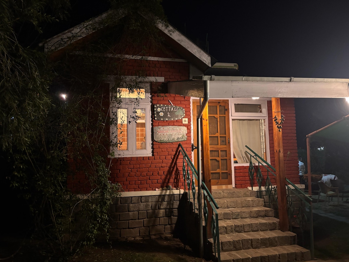 The cottage is viewed at night, featuring a red brick exterior and illuminated windows. A welcoming porch is accessed by a set of stone steps, and a wooden sign indicating the cottage name hangs prominently. Surrounding trees provide a natural backdrop.