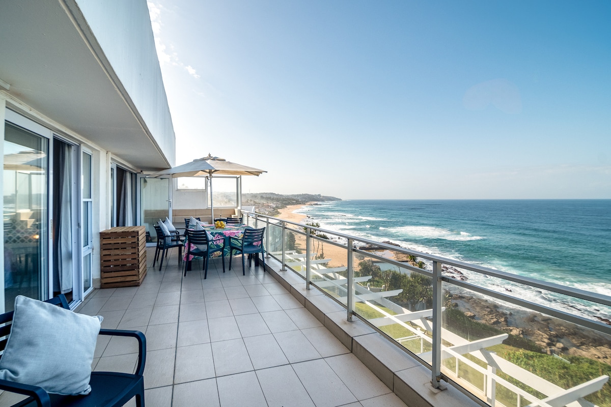 A private balcony features a dining table and chairs, providing an unobstructed view of the ocean and coastline. Sun umbrellas are positioned to offer shade. The surrounding area displays tiled flooring, enhancing the space's open and airy feel, with the blue sky complementing the serene sea.