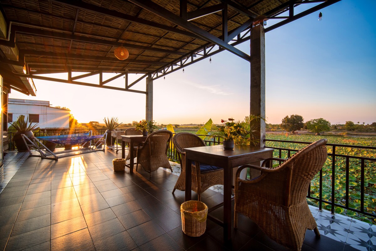 A spacious patio area features elegant wooden tables and comfortable wicker chairs, providing a setup for outdoor dining. Lush lotus farms stretch into the distance, illuminated by soft sunset light, creating a serene backdrop for relaxation.