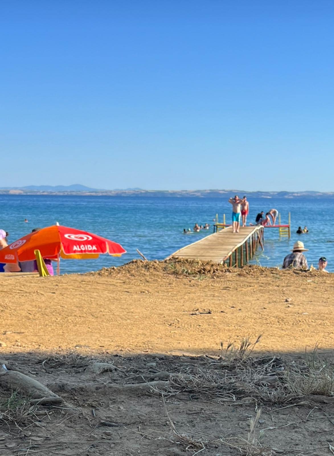 A sandy beach extends towards calm waters, where a wooden dock leads into the sea. Brightly colored umbrellas provide shade for beachgoers, while several people are seen enjoying the water. The horizon features gentle waves and distant hills, under a clear blue sky.