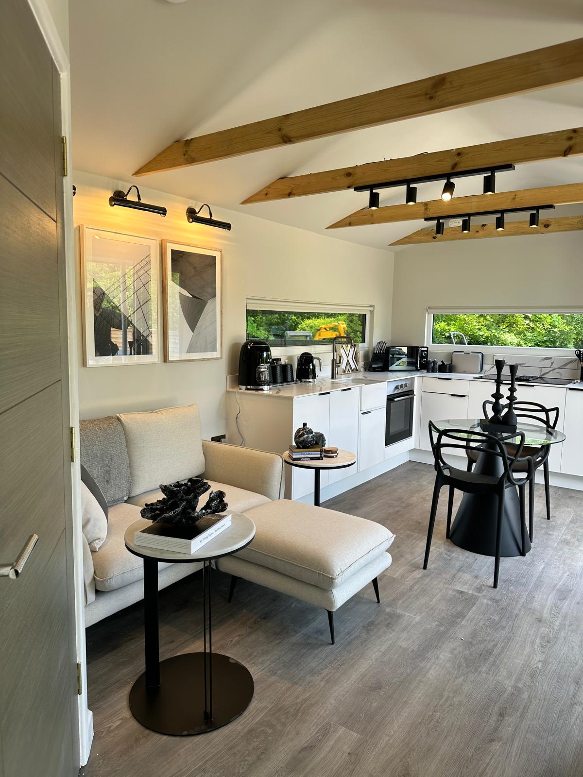 A modern living space features light wood beams overhead and a stylish seating area with a light-colored sofa and a small side table. Black dining chairs surround a table in the background, with kitchen appliances visible through a large window overlooking greenery.
