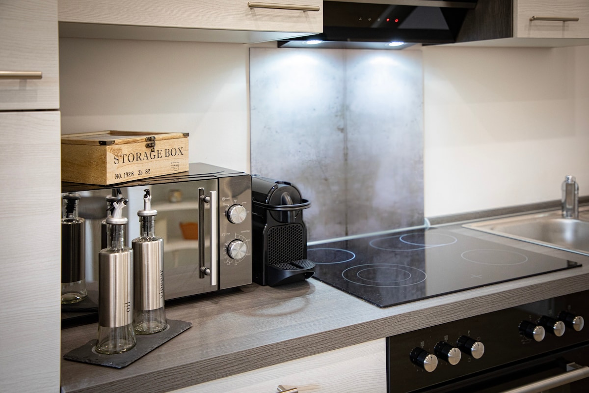 A modern kitchen area is featured, showcasing sleek cabinetry with a neutral finish. Countertops host a microwave, a Nespresso machine, and stylish storage containers. A hob with four cooking zones is visible, along with a stainless-steel sink, creating an inviting cooking space.