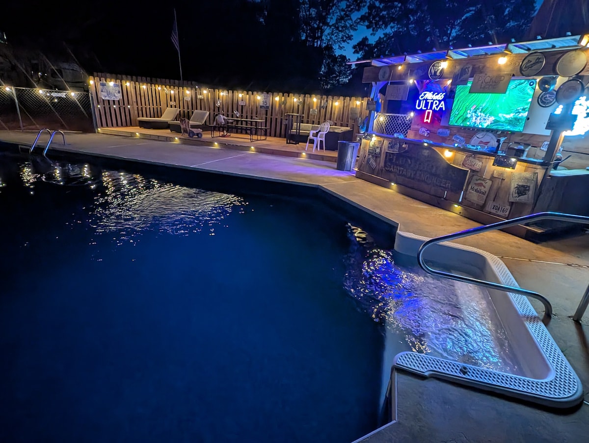 The inviting pool area is presented under soft ambient lighting, with a bar setup visible in the background. Lounge chairs line the poolside, complementing the tranquil water that reflects the surrounding lights, creating a serene atmosphere for relaxation.