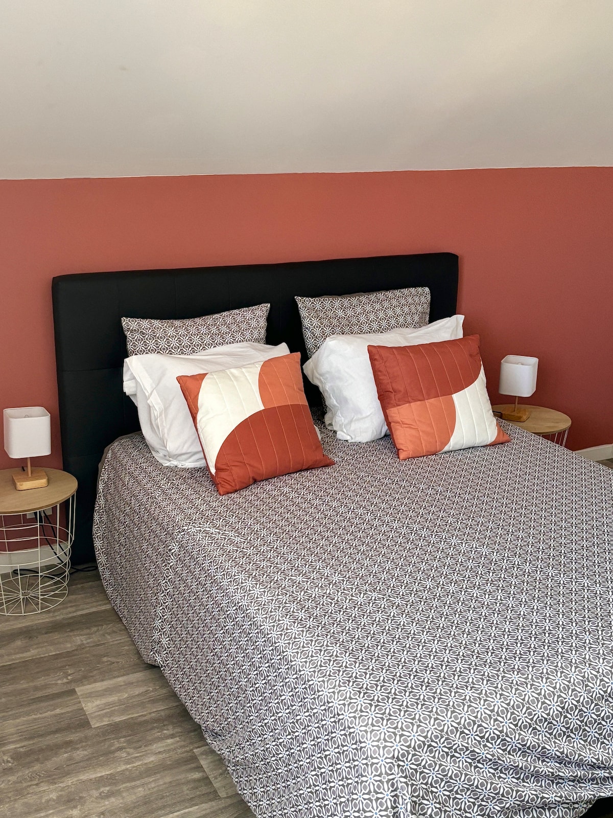 A comfortable sleeping area features a black headboard with two pillows in complementary colors. The bed is dressed with a patterned duvet cover, and two bedside lamps are placed on woven side tables. Light streams in from an unseen source, enhancing the restful ambiance.