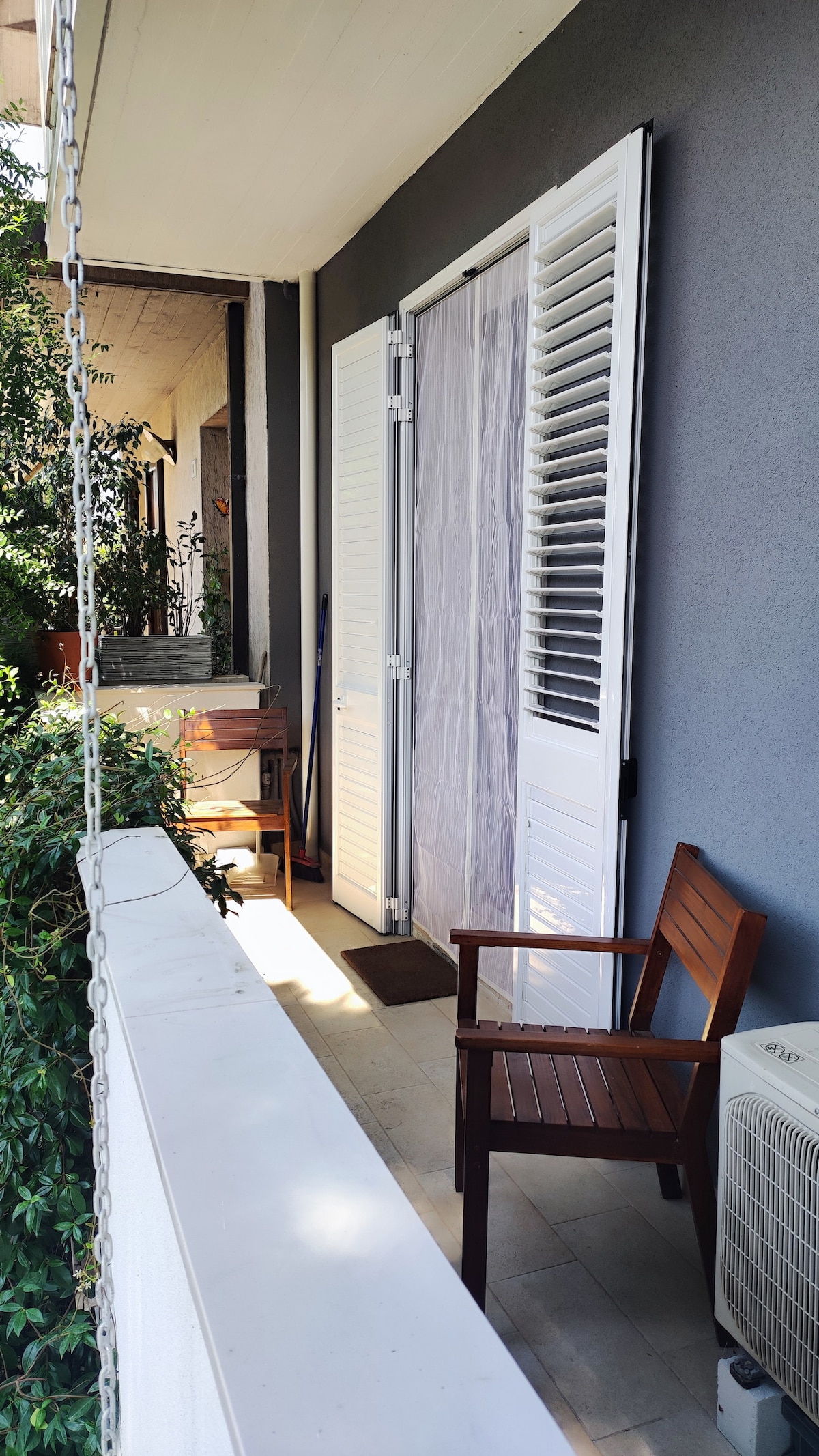A small balcony is featured with a wooden chair positioned by a partially open white door. Lush greenery surrounds the space, enhancing privacy and a sense of tranquility. A compact air conditioning unit is visible to the side, along with a natural stone floor.
