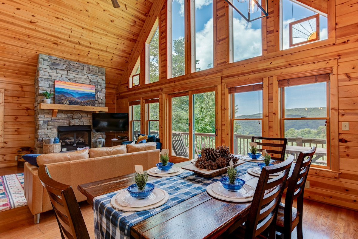 The open living area features a high wood-paneled ceiling, floor-to-ceiling windows, and a cozy stone fireplace. A wooden dining table is set with blue plates and centerpieces, while a sectional couch provides comfort. Scenic mountain views are framed by the bright sunlight streaming in.