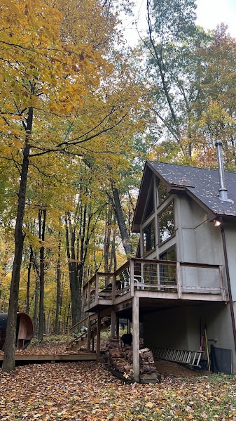 A multi-level cabin is surrounded by towering trees adorned in autumn leaves. The structure features a spacious deck with railing, providing outdoor seating. A wood-fired hot tub is visible nearby, offering a relaxing spot amidst the natural landscape.