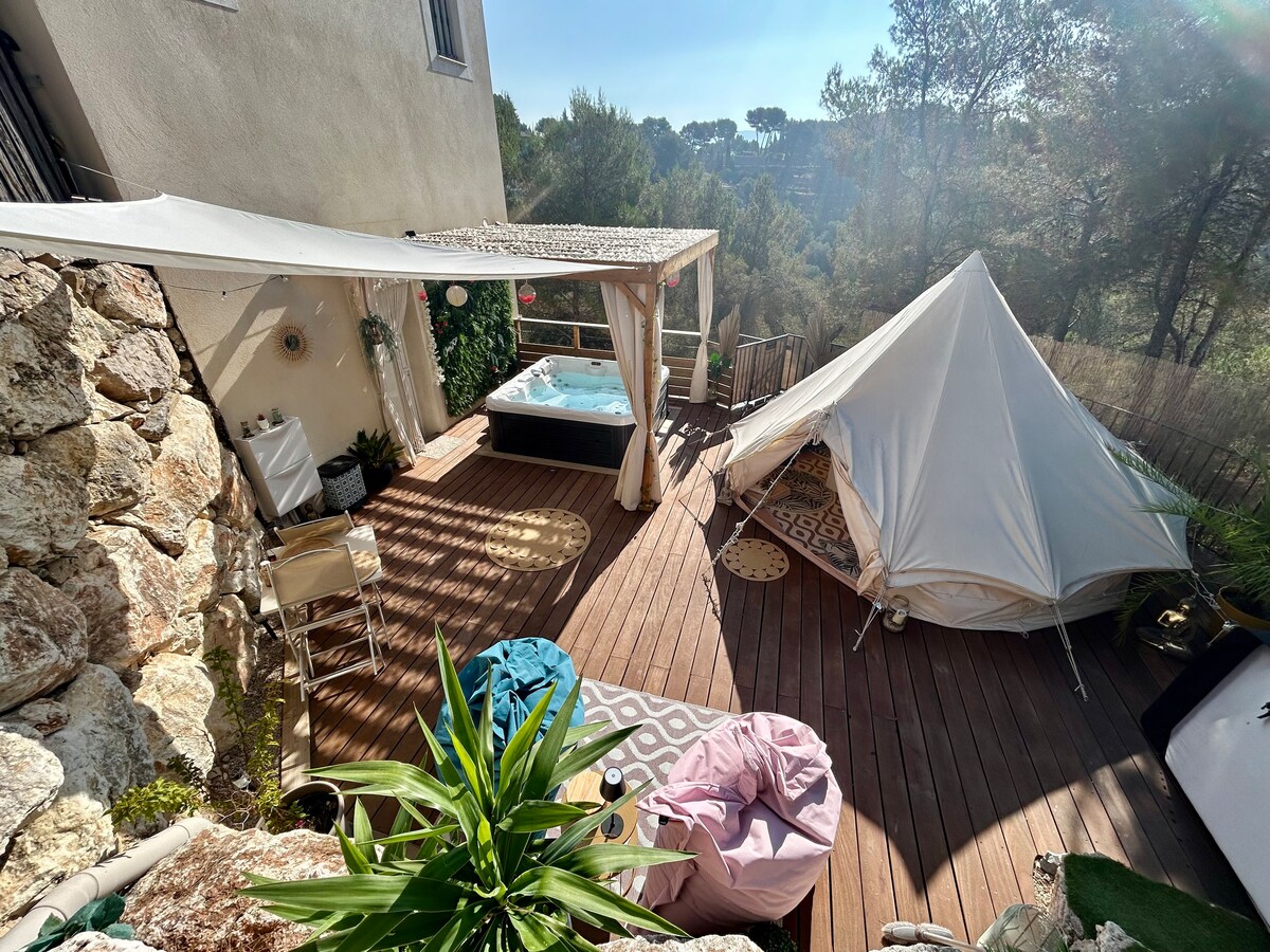 A spacious outdoor terrace features a private Jacuzzi, surrounded by lush greenery. A white tent lodge is positioned nearby on wooden decking, accompanied by colorful poufs and a small table. Sunlight illuminates the area, enhancing the natural beauty of the setting.