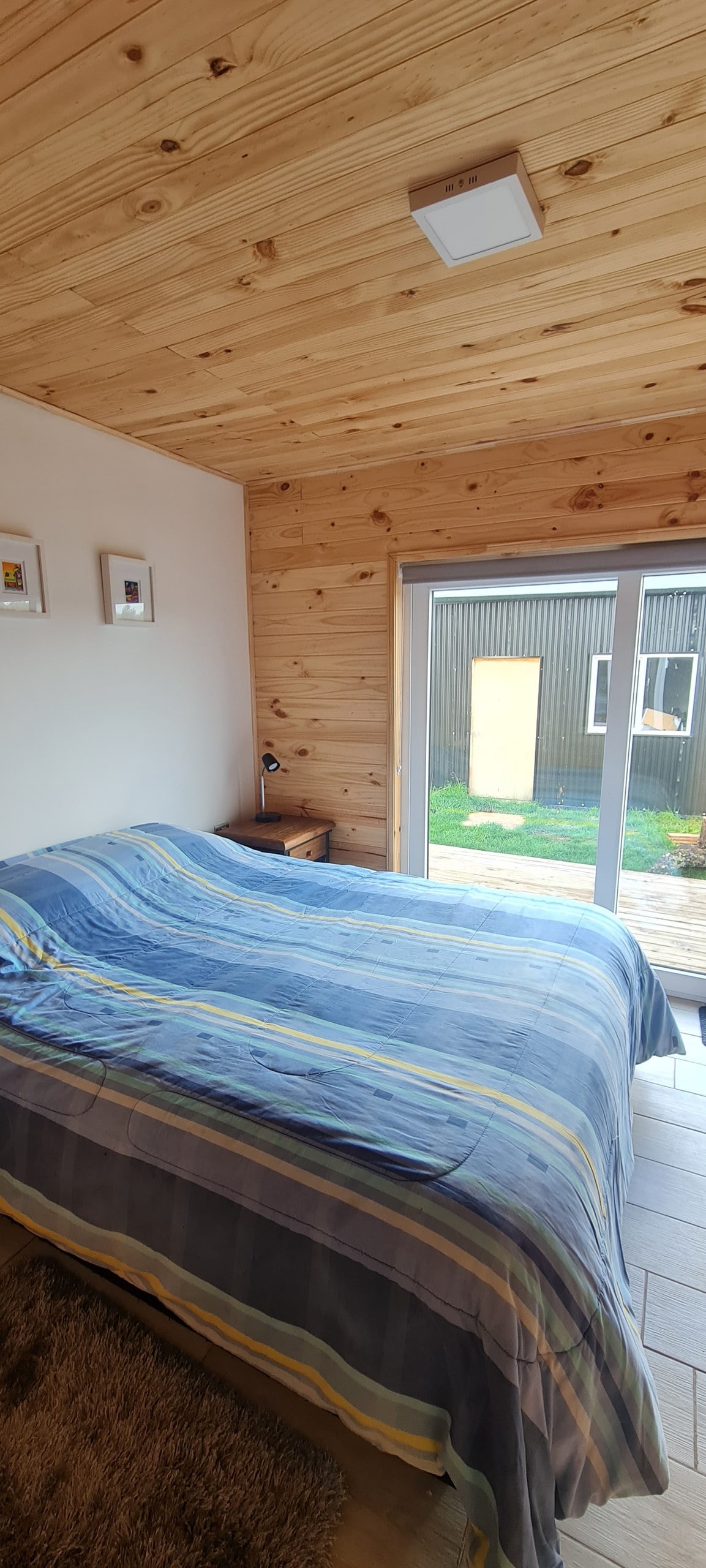 The bedroom features a spacious bed with a striped blanket, positioned beside a small nightstand with a lamp. Large sliding glass doors allow natural light to enter, leading to a wooden deck and views of the surrounding outdoor space.