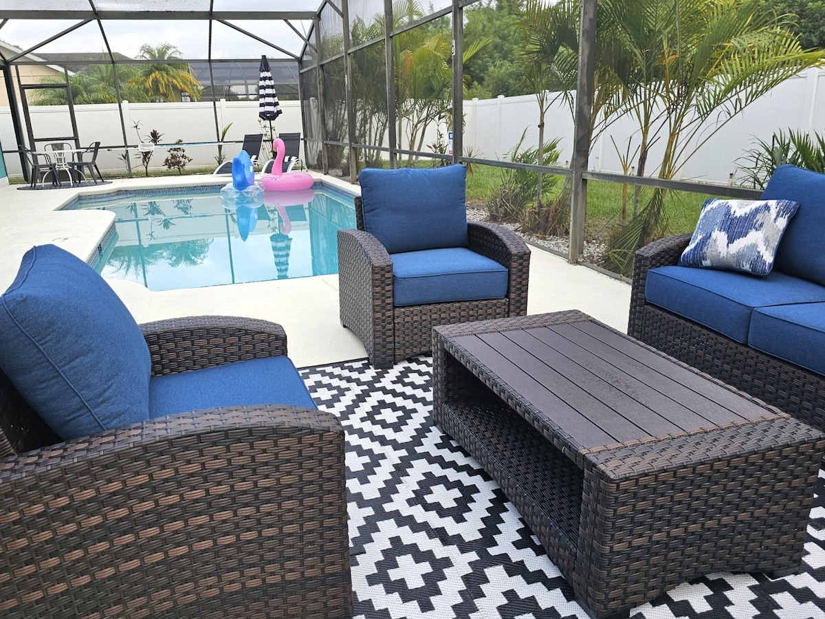 An outdoor patio area is showcased, featuring comfortable seating with blue cushions arranged around a low table. A geometric rug is laid on the floor, and the fenced-in pool area is visible, complete with floating pool toys and surrounding greenery.