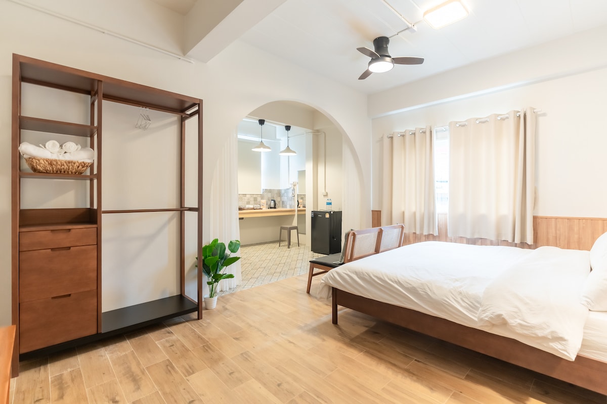 A simple and modern room features a spacious bed with fresh linens. A wooden wardrobe and a small potted plant add to the decor. Light-colored curtains create a bright atmosphere, while an open archway leads to the kitchenette area visible in the background.